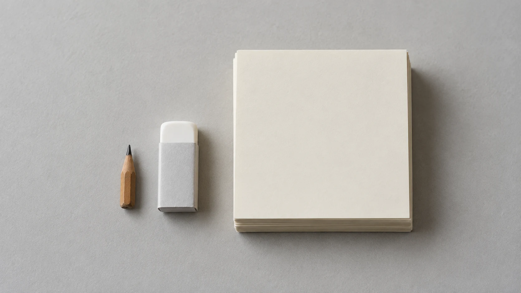An editorial photograph looking straight down on a pale grey surface. A pencil worn to a short stub and an unused white eraser still in its paper sleeve sit beside a squared stack of blank cream sheets.