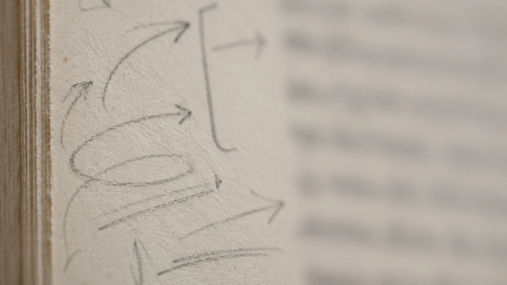 An editorial photograph of the margin of an old book page in extreme close-up. Faint graphite pencil arrows, brackets, loops and underlines crowd the cream paper, while the printed column beside them dissolves into an unreadable blur.