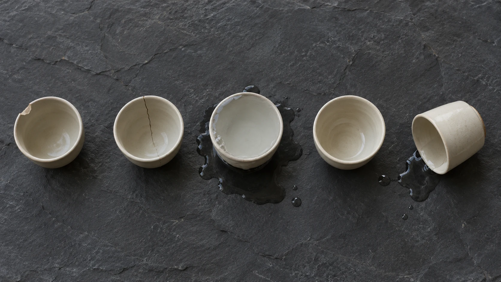 An editorial photograph, shot from directly above, of five cream ceramic cups in a row on dark grey slate, each failing differently. One has a chipped rim, one a hairline crack, one brims over into a puddle, one sits empty, and the last lies tipped on its side beside a small spill.