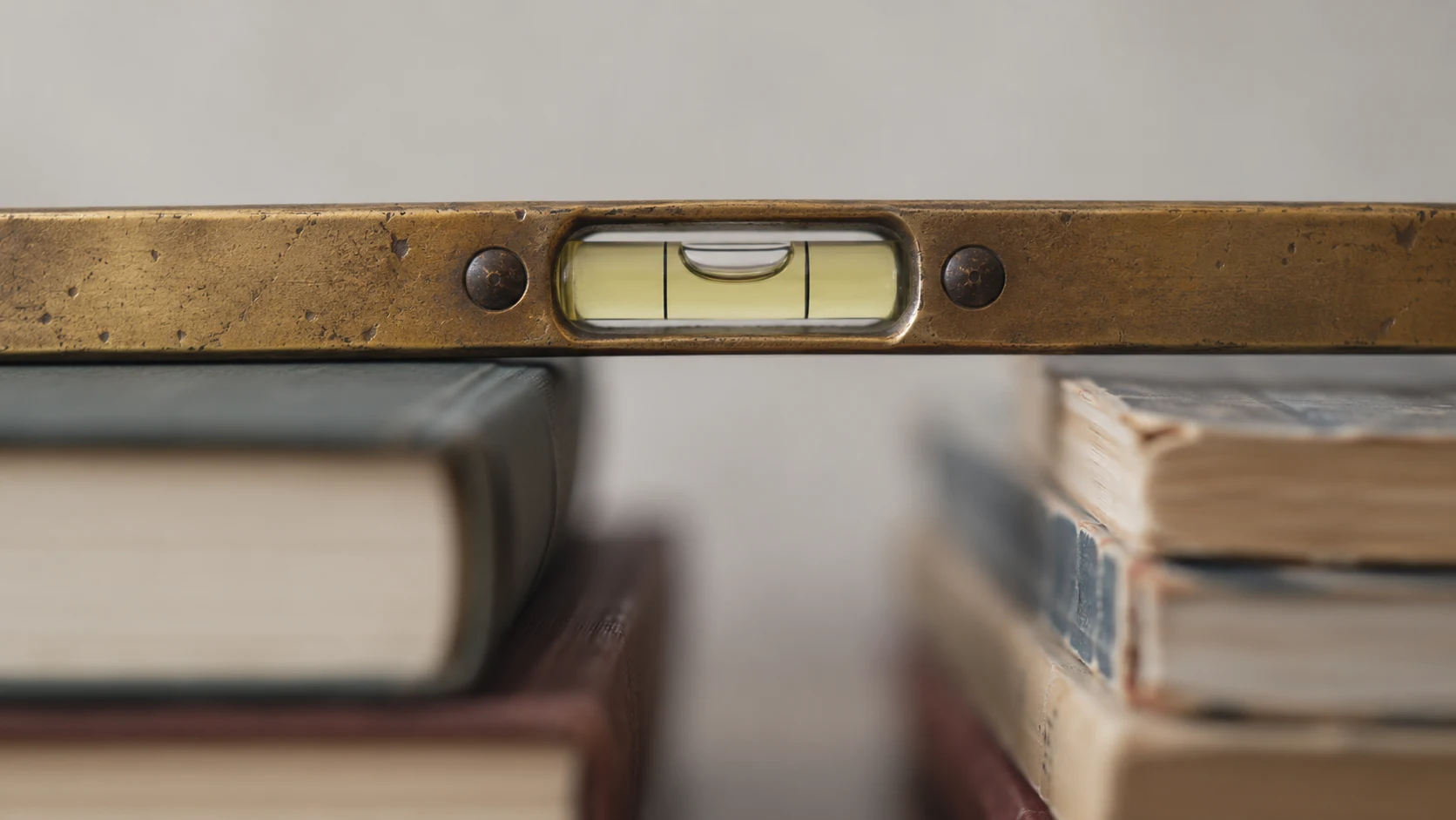 An editorial photograph of a worn brass spirit level in close-up, its bubble resting dead centre. The level lies across two very different stacks of books, thick cloth hardbacks on one side and frayed old paperbacks on the other, both softly blurred.
