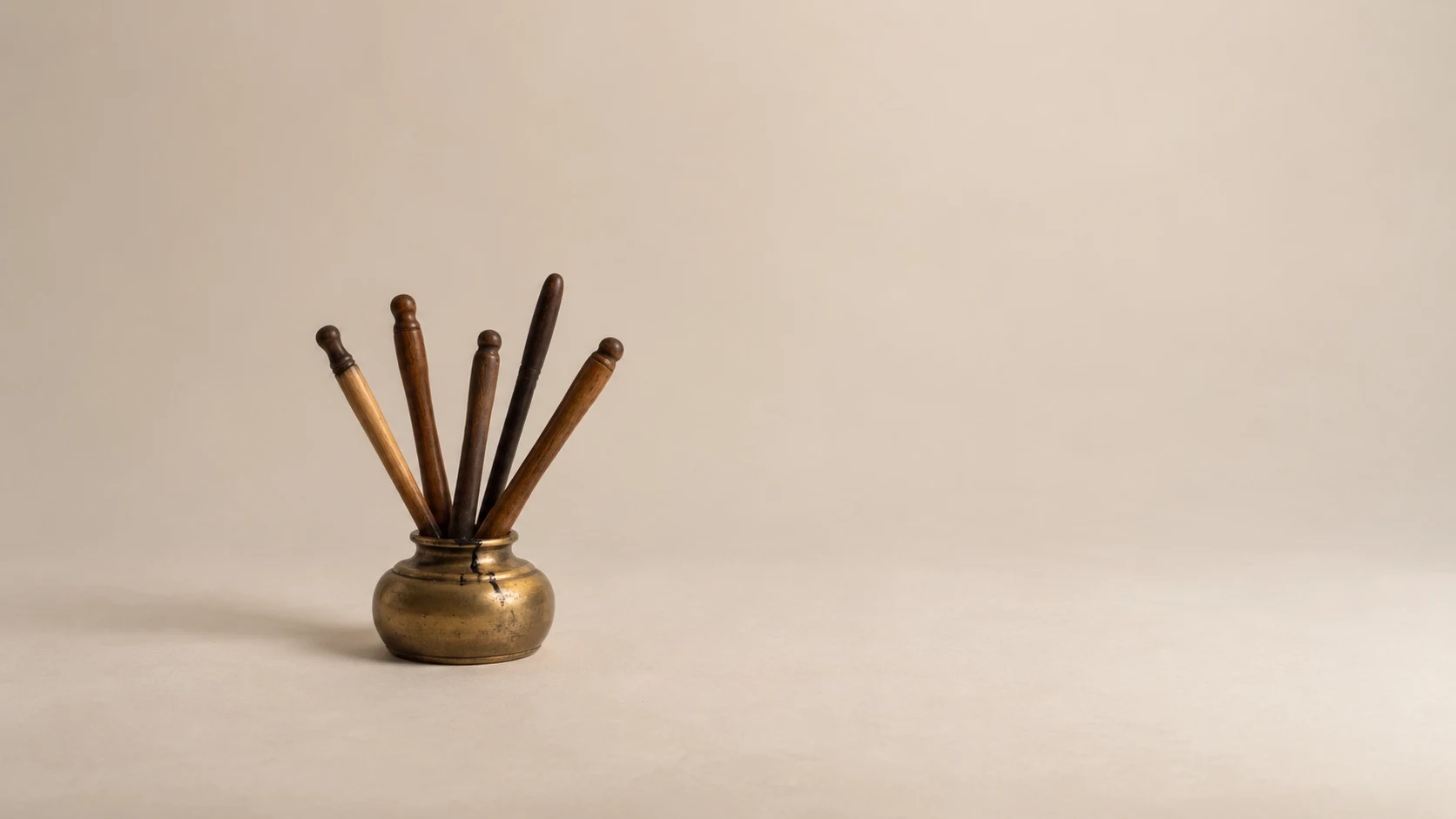 An editorial photograph of a small squat brass inkwell on a seamless cream backdrop, with five wooden dip pens crowded into its single narrow mouth, nibs down in the ink. A thin trickle of black ink runs down the rim, and the rest of the frame is empty.