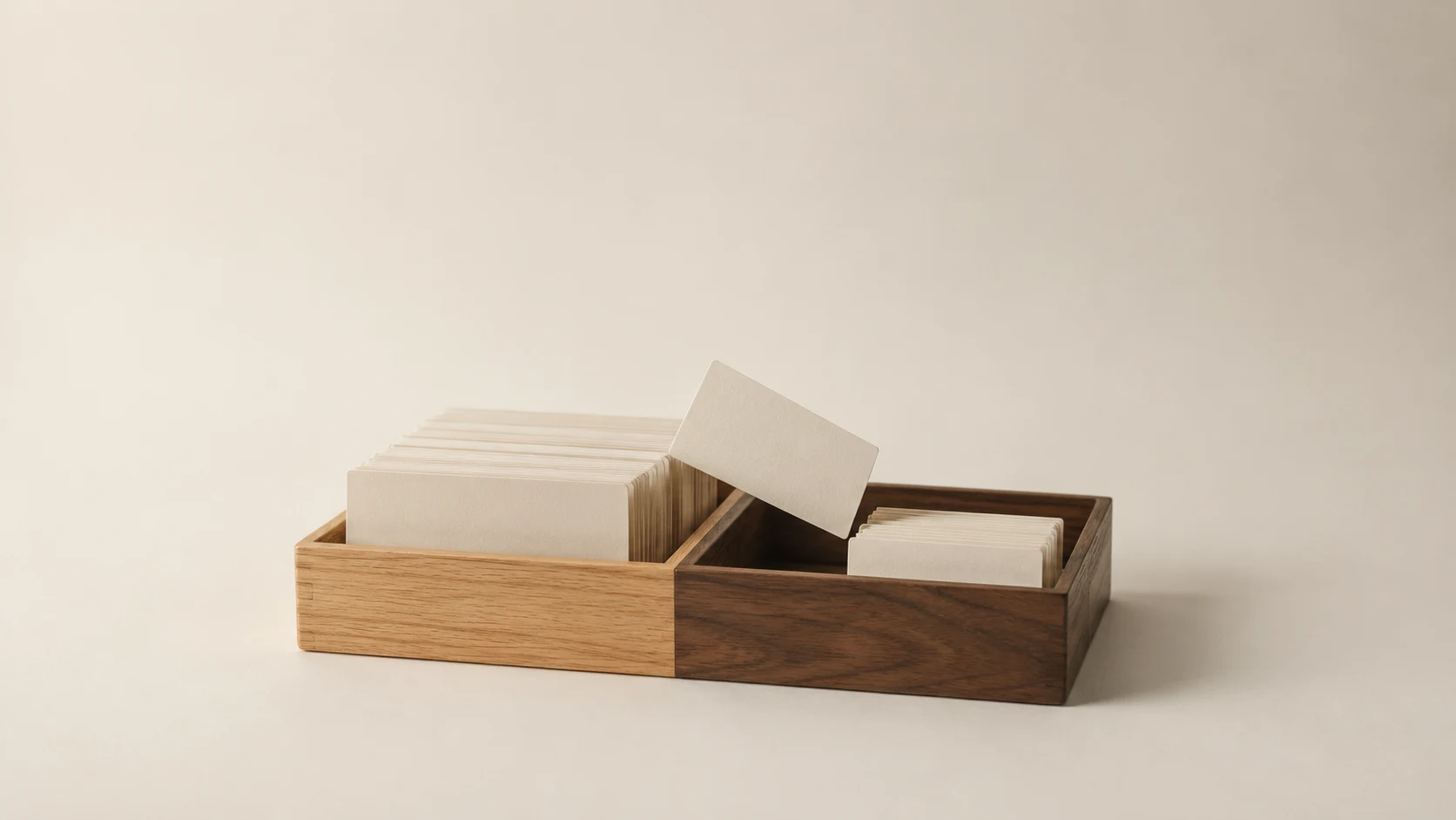 An editorial photograph of a two-part sorting tray on a seamless cream backdrop, one half pale oak and the other walnut. The oak side is packed with blank cream cards, the walnut side holds only a few, and one card leans across the divider mid-sort.