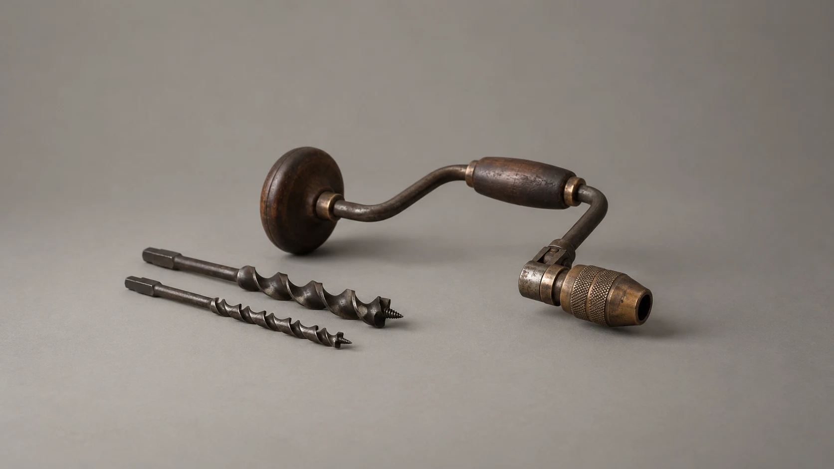 An editorial photograph of an old hand brace drill with a wooden handle and an empty brass chuck, lying on a seamless warm-grey backdrop. Two interchangeable auger bits rest beside it.