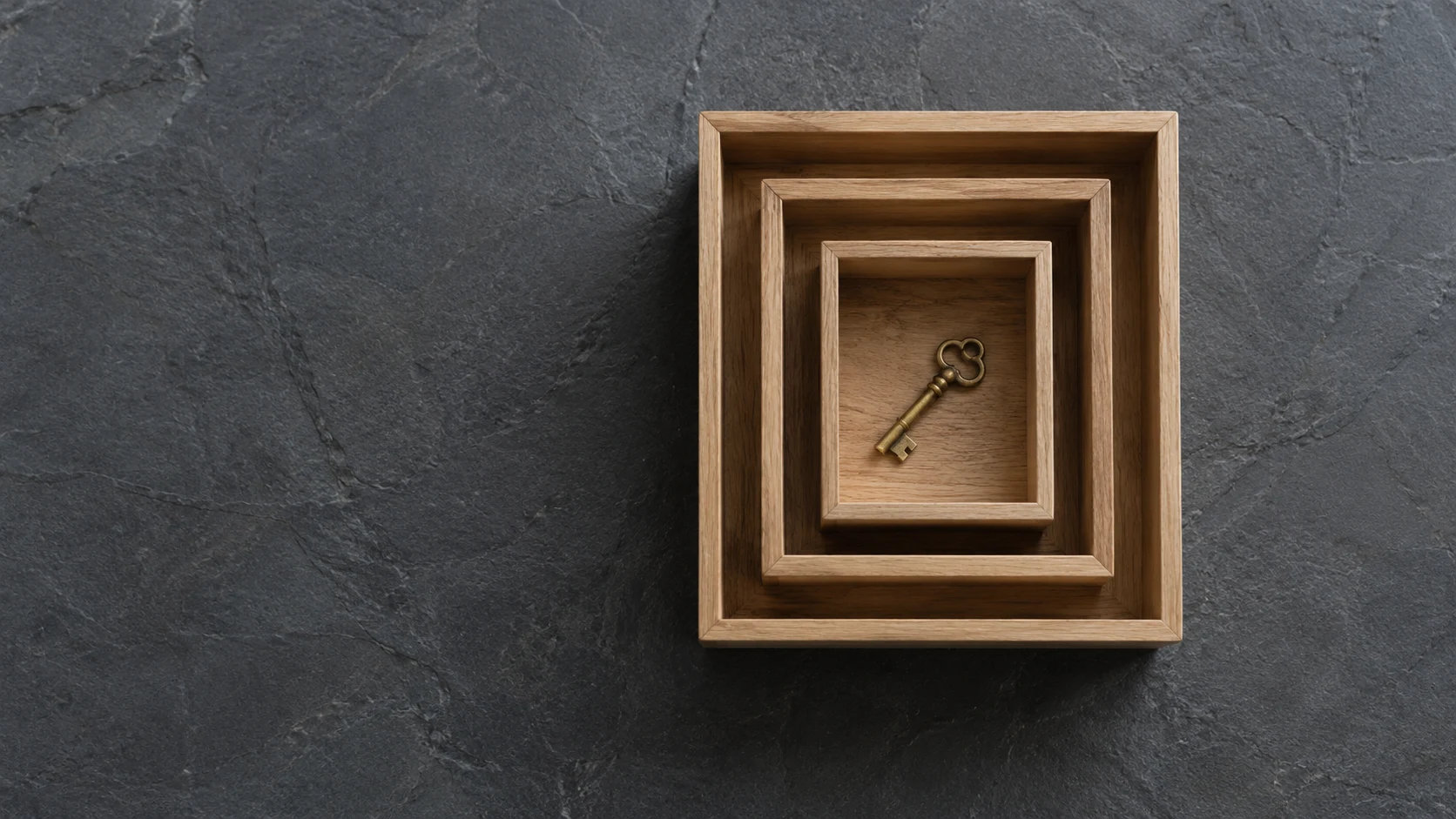 An editorial photograph of three lidless light oak boxes nested one inside another, seen from directly above on dark grey slate. A small brass key lies in the innermost box.
